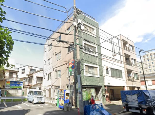 - 繁華街にある緑色の外壁を持つ集合住宅、近くに駐車場と自動販売機、青空が広がるシーン