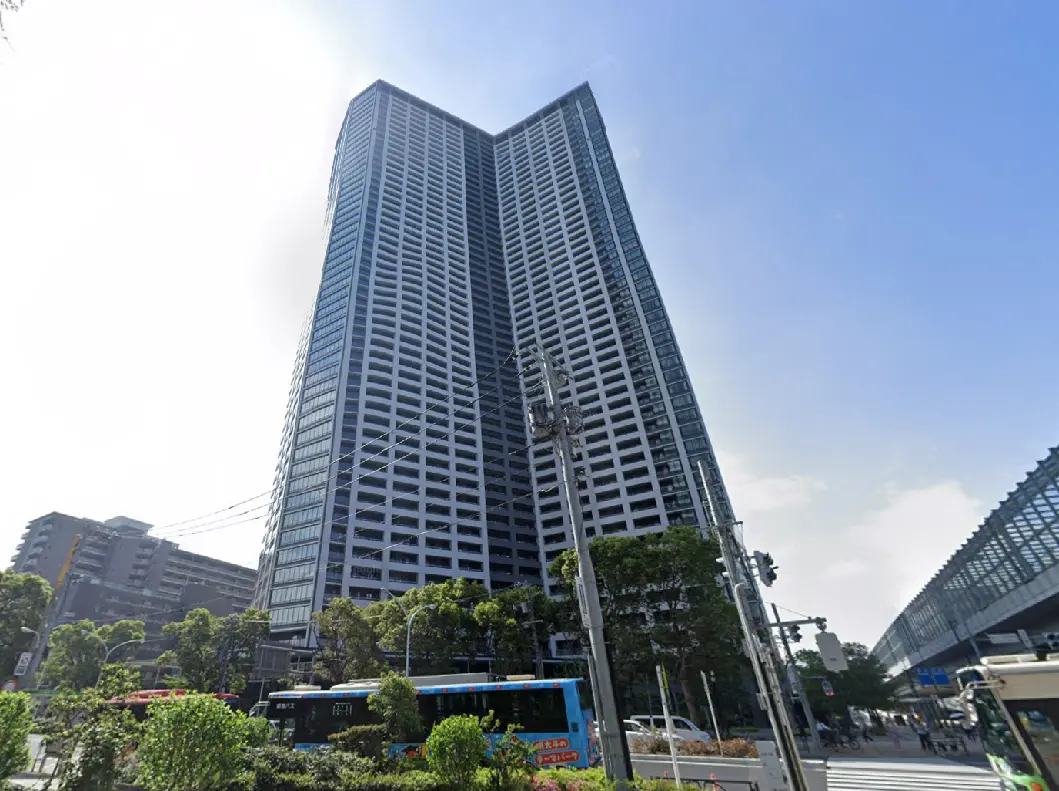 - 高層ビルの外観、周囲には緑豊かな街並みとバス停、青空が広がる風景