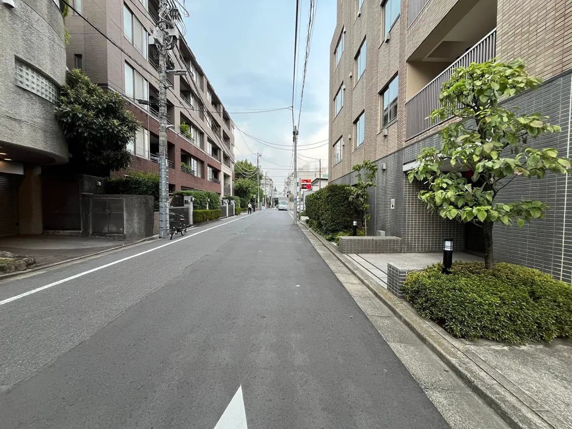 - 都会的な住宅街の通り、左右に並ぶ赤茶色のマンションと緑豊かな植栽、穏やかな雰囲気の景観
