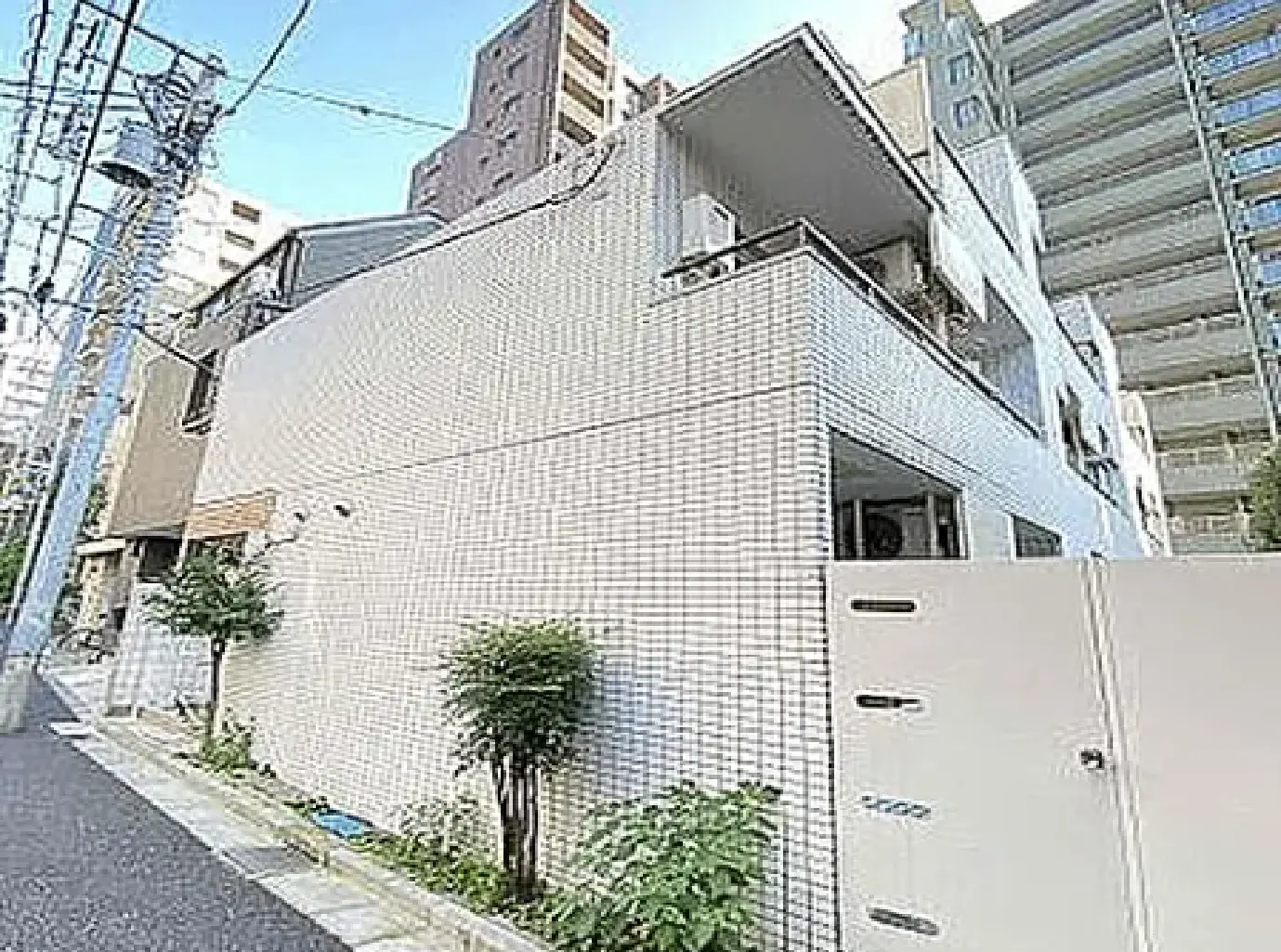 - ホワイトタイルで覆われた現代的な建物の外観、周囲に緑の植栽と近隣の高層マンションが見える街並み