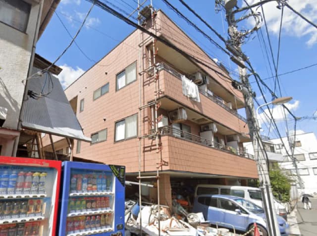 - 鉄筋コンクリート製の茶色い外壁のマンション、前景には自動販売機と数台の車、周囲には電線が張り巡らされている街並みの風景