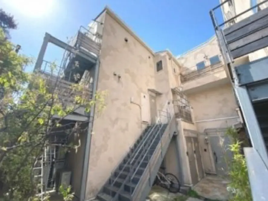 - 明るい青空の下、外階段とバルコニーを持つビルの側面、周囲には緑の植物が見える景観