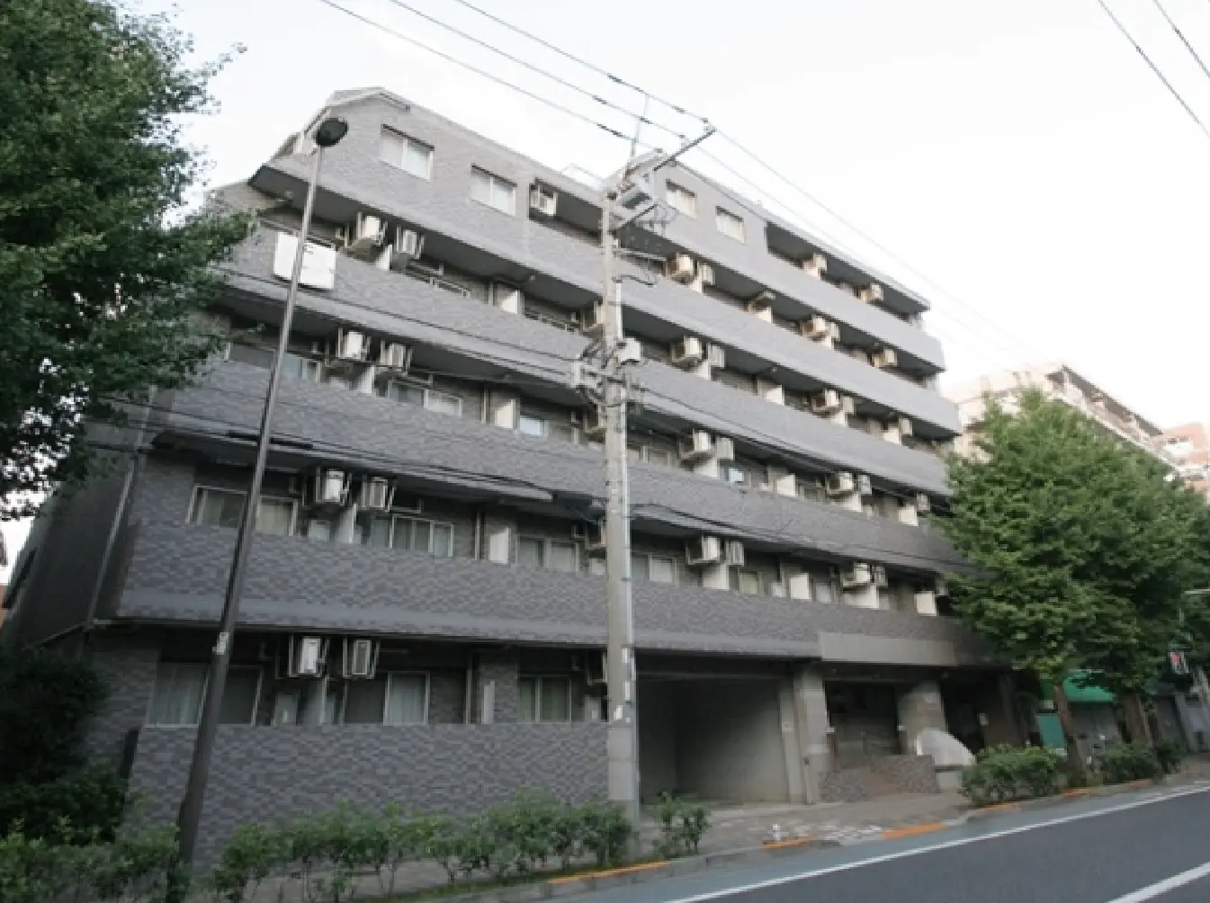 - グレーの外観を持つ5階建てのアパートメントビル、周囲には緑の木々と街路灯、エアコンが設置された窓が目立つ外観