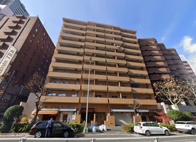 - 都市型マンションの外観、7階建てのレンガ調のファサード、隣接する建物と青空の下の景観