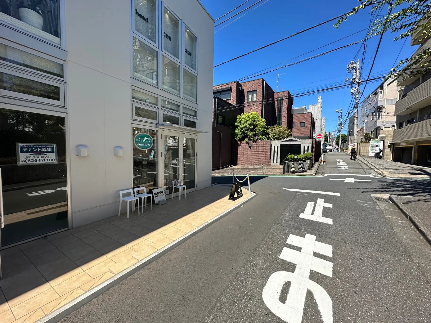 白い外壁の現代的なビルとその前の道路、歩道にテーブルと椅子が置かれたカフェが見える都市の風景