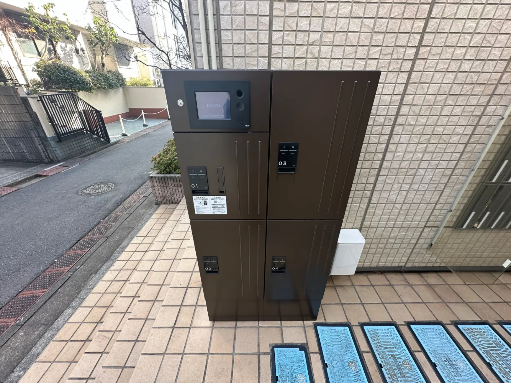 マンションのエントランスに設置された宅配ボックス、D-roomロゴ付き、タッチパネル操作、街路と背景の建物が見える外観