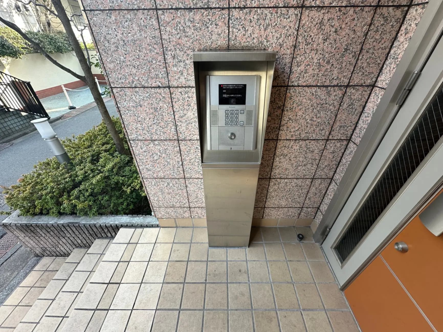 ビルの入り口に設置されたインターホン、タイル張りの床と壁、緑の植え込みが見える外観