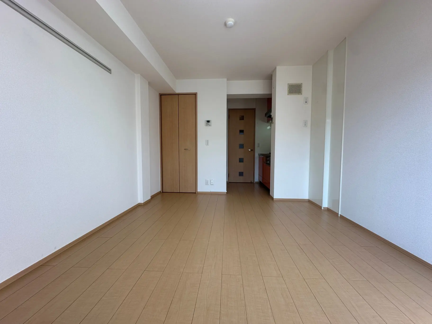 空の部屋、白い壁、茶色の木製フローリング、キッチンへの扉が見えるインテリア