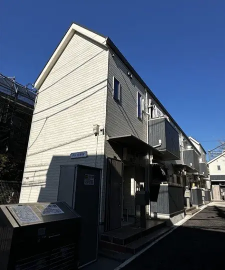 - 明るい空の下に立つ二階建てのアパートメント、白い外壁と灰色のバルコニー、周囲には建物と設備が見える街並み