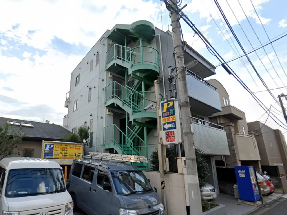 - メタリックな緑色の螺旋階段が特徴的なアパート外観、駐車場の案内看板と並んだ車両が視認できる通りの風景