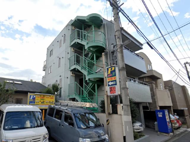 - メタリックな緑色の螺旋階段が特徴的なアパート外観、駐車場の案内看板と並んだ車両が視認できる通りの風景