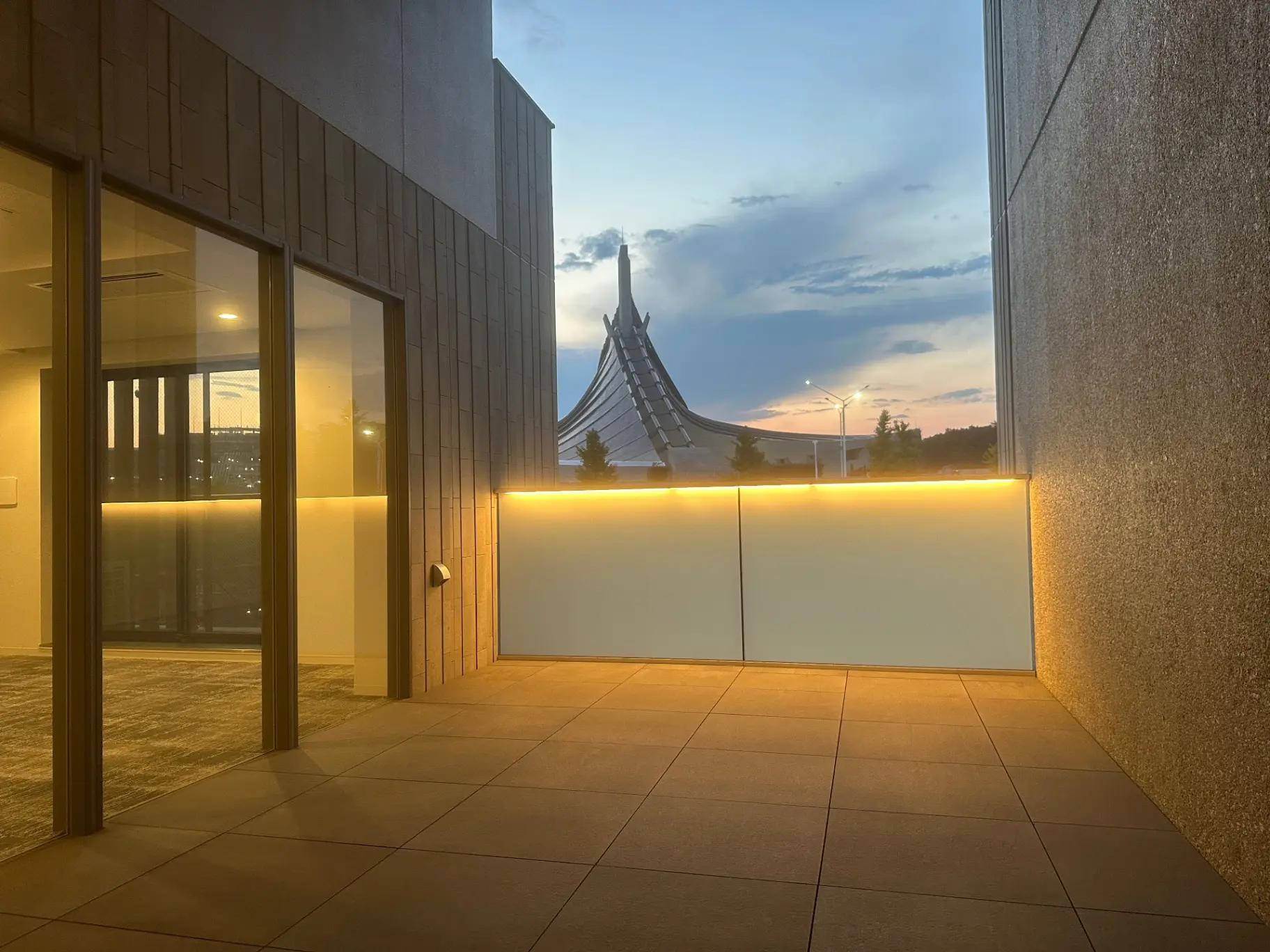 モダンな外観のビルのエントランス、壁にはイエローのライトが照らされ、背景には夕暮れ時の空と建築物のシルエット