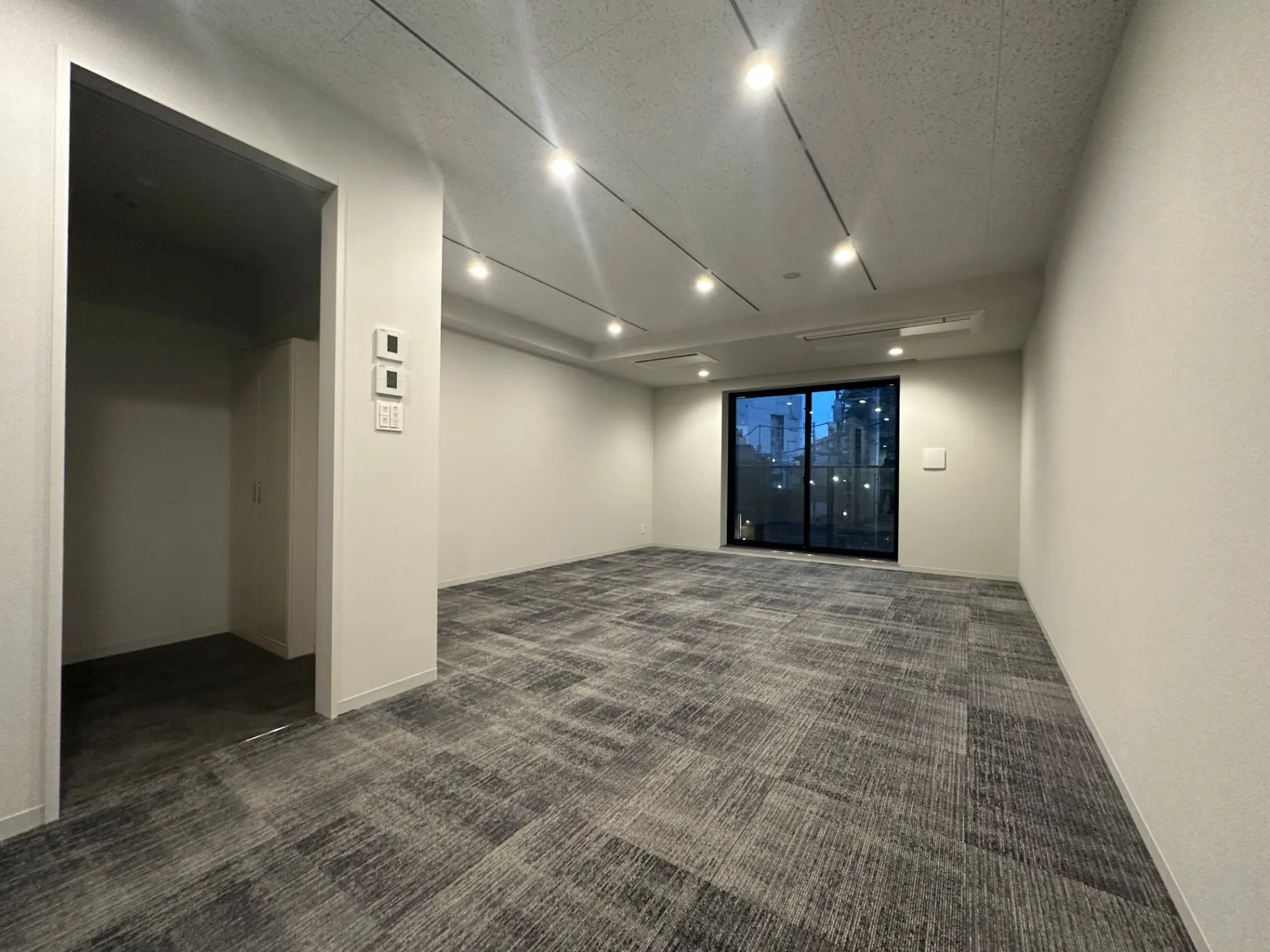 広々としたオフィススペース、グレーのカーペットフロア、多数の天井照明、ガラスの掃き出し窓とクリアな壁