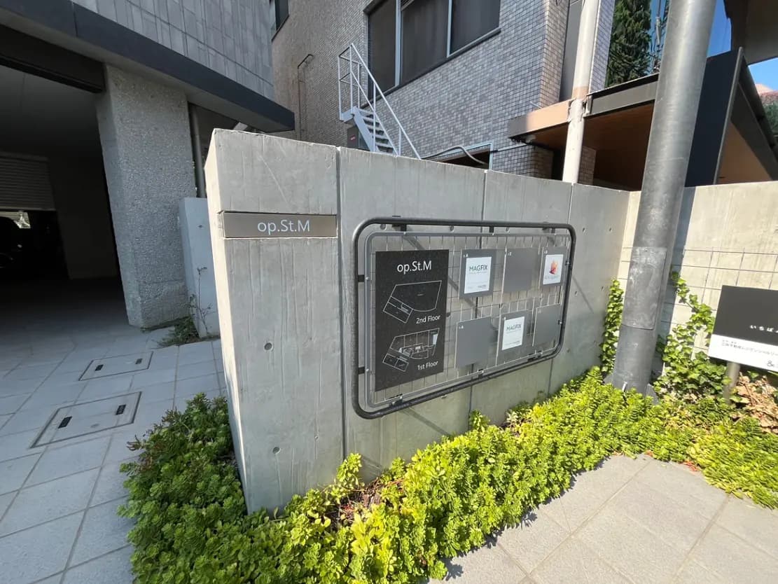 - モダンな建物の入り口、コンクリート製の看板に「op.St.M」と記載、周囲に緑の植物が配されている景観