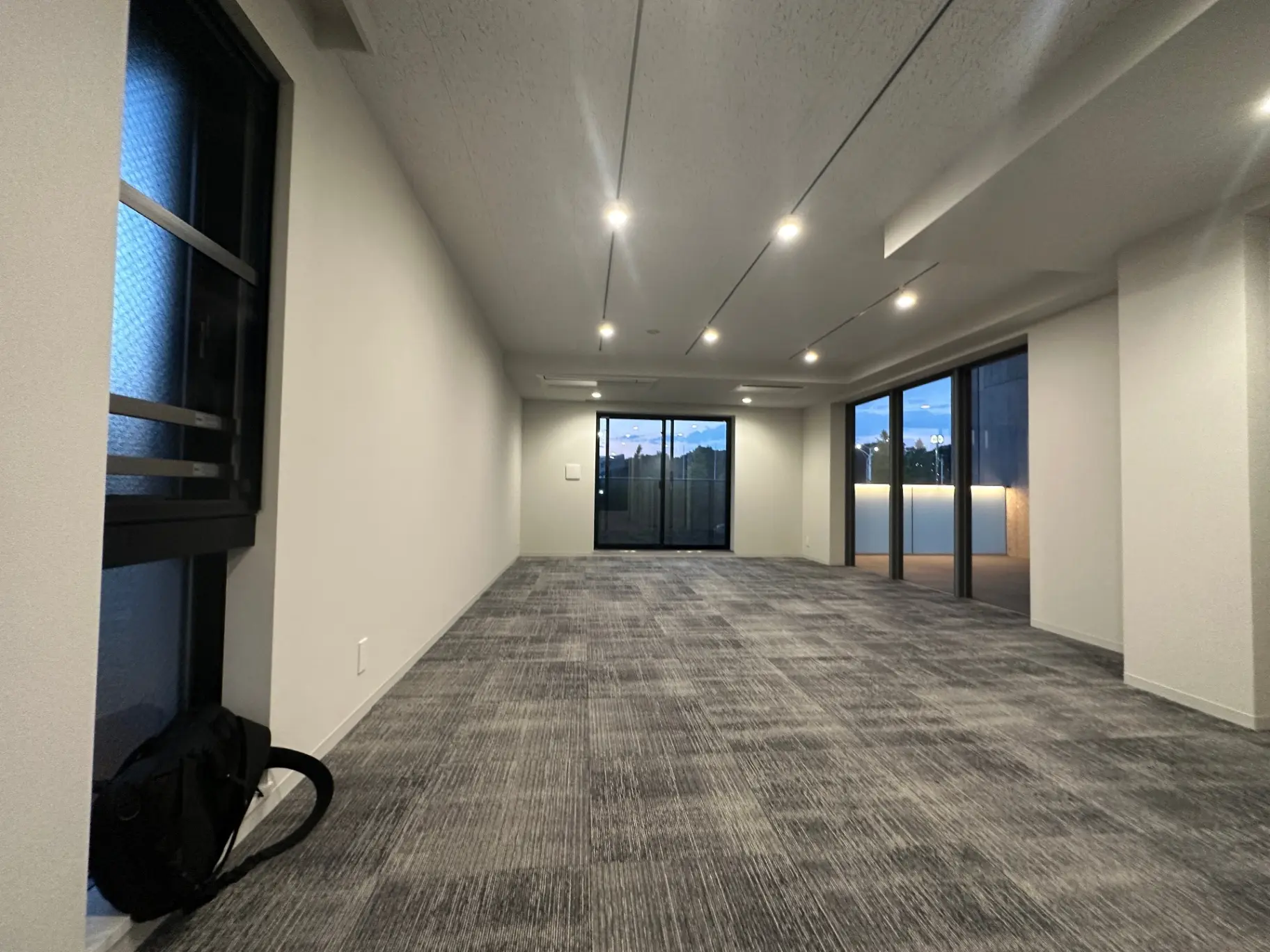 空間が広いモダンなオフィスの空間、ダークグレーのカーペットと白い壁、大きな窓とLED照明が特徴的なインテリア