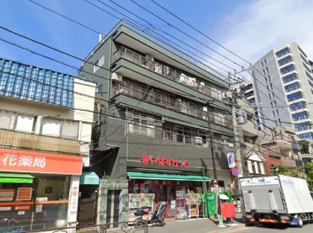 - 都市の風景に立つ住宅ビルと花屋、赤い看板が目立つ店舗の外観、青空と電線が広がる明るい昼間の風景