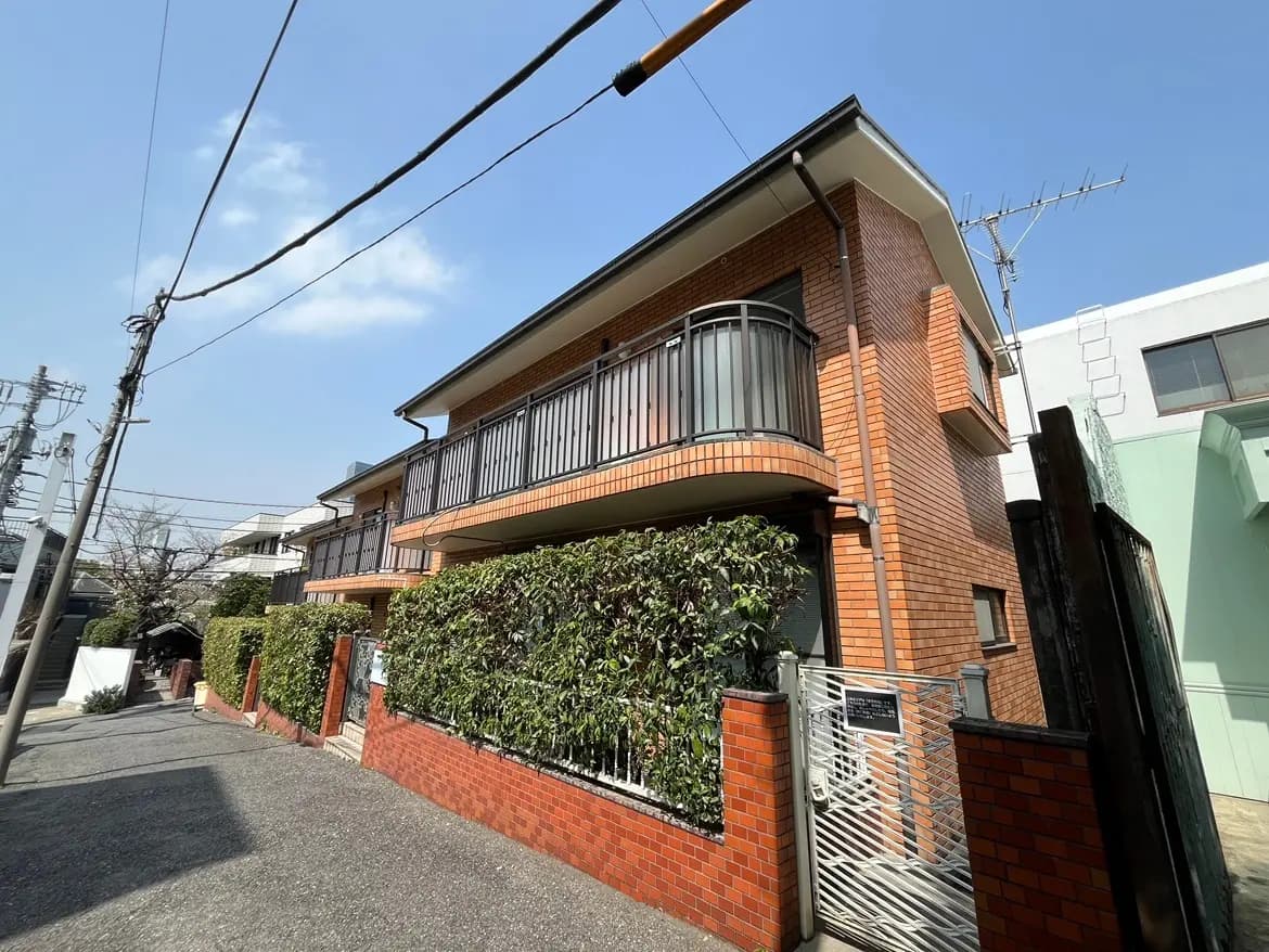 晴れた日の住宅街にあるレンガ造りの２階建ての家、バルコニーと緑豊かな生け垣がある外観