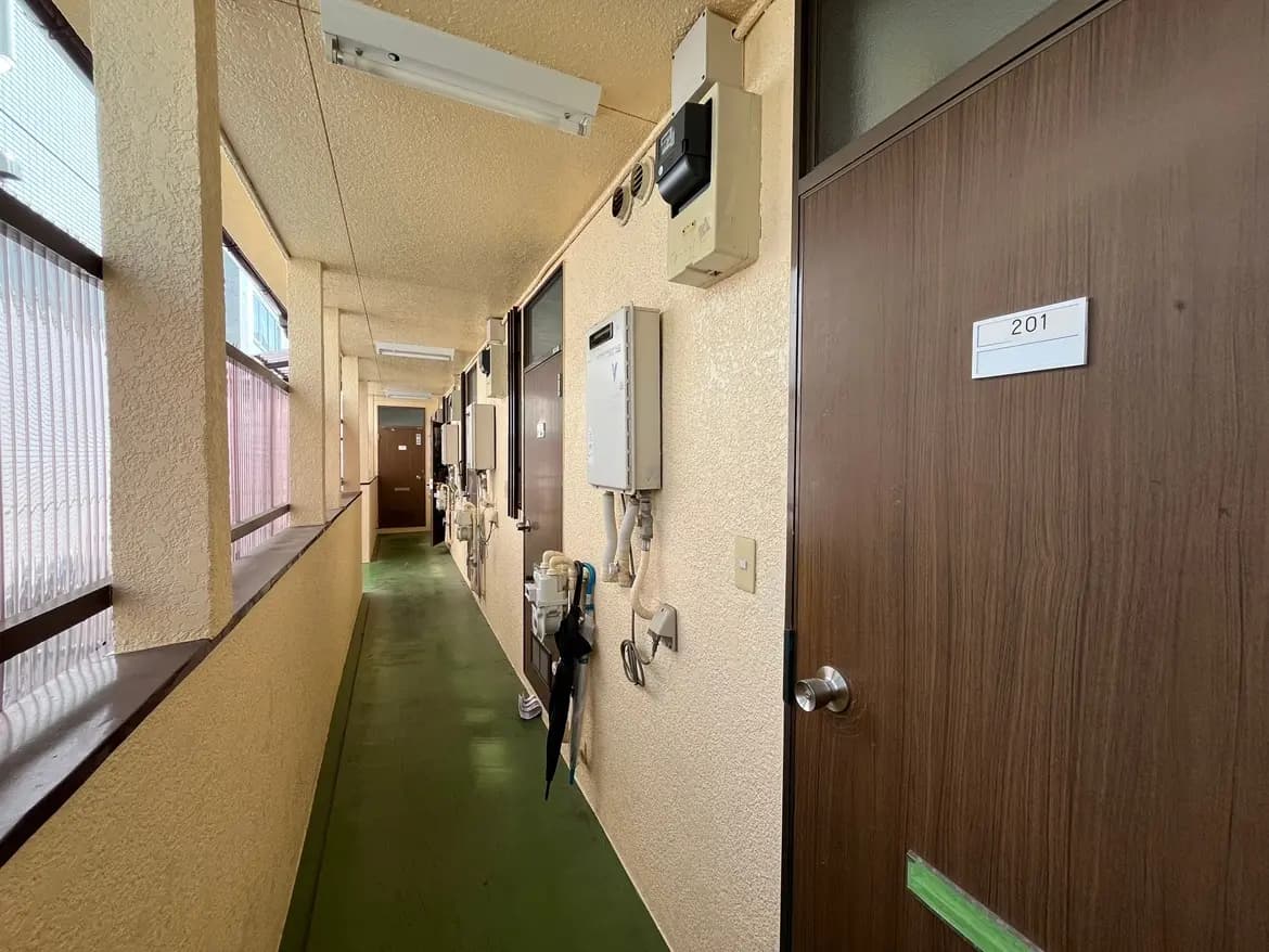 狭いアパートの廊下、左側に窓と掲示板、右側に部屋のドア201号室があるインテリア