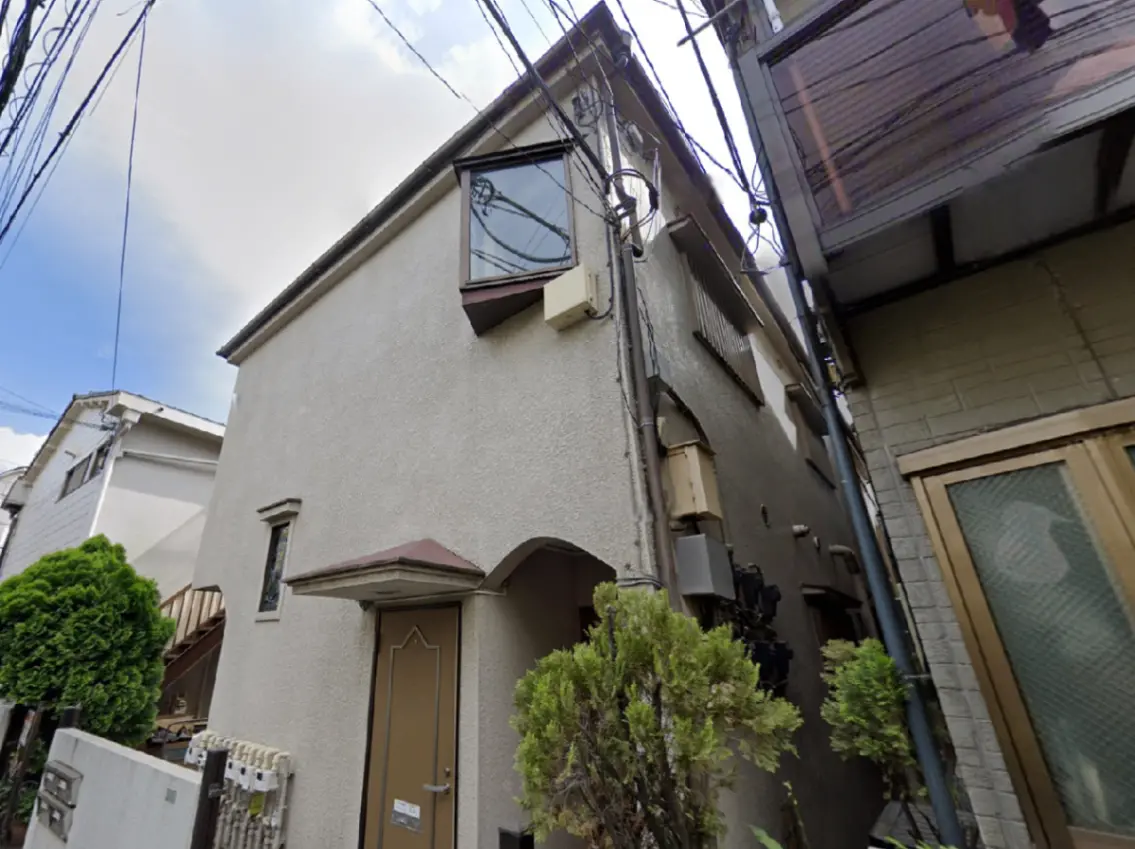 - 狭い路地に建つシンプルな外観の住宅、玄関ドアと屋根付き入口が特徴的で周囲に緑がある風景