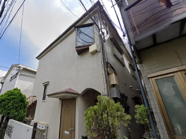 - 狭い路地に建つシンプルな外観の住宅、玄関ドアと屋根付き入口が特徴的で周囲に緑がある風景