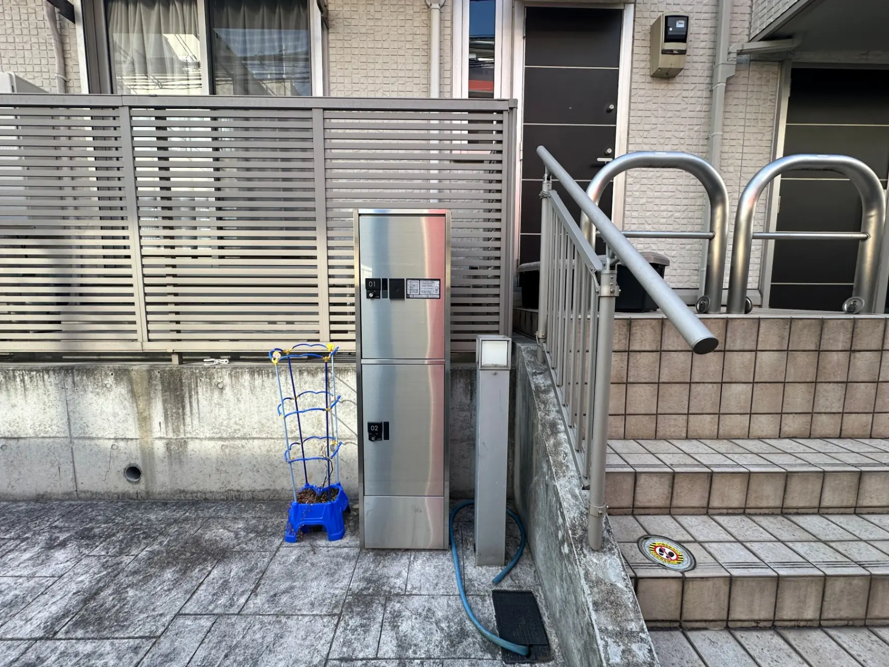 住宅街の玄関先、スリッパラック、手すり付き階段、宅配ロッカー、防犯カメラが設置されたエントリーエリア