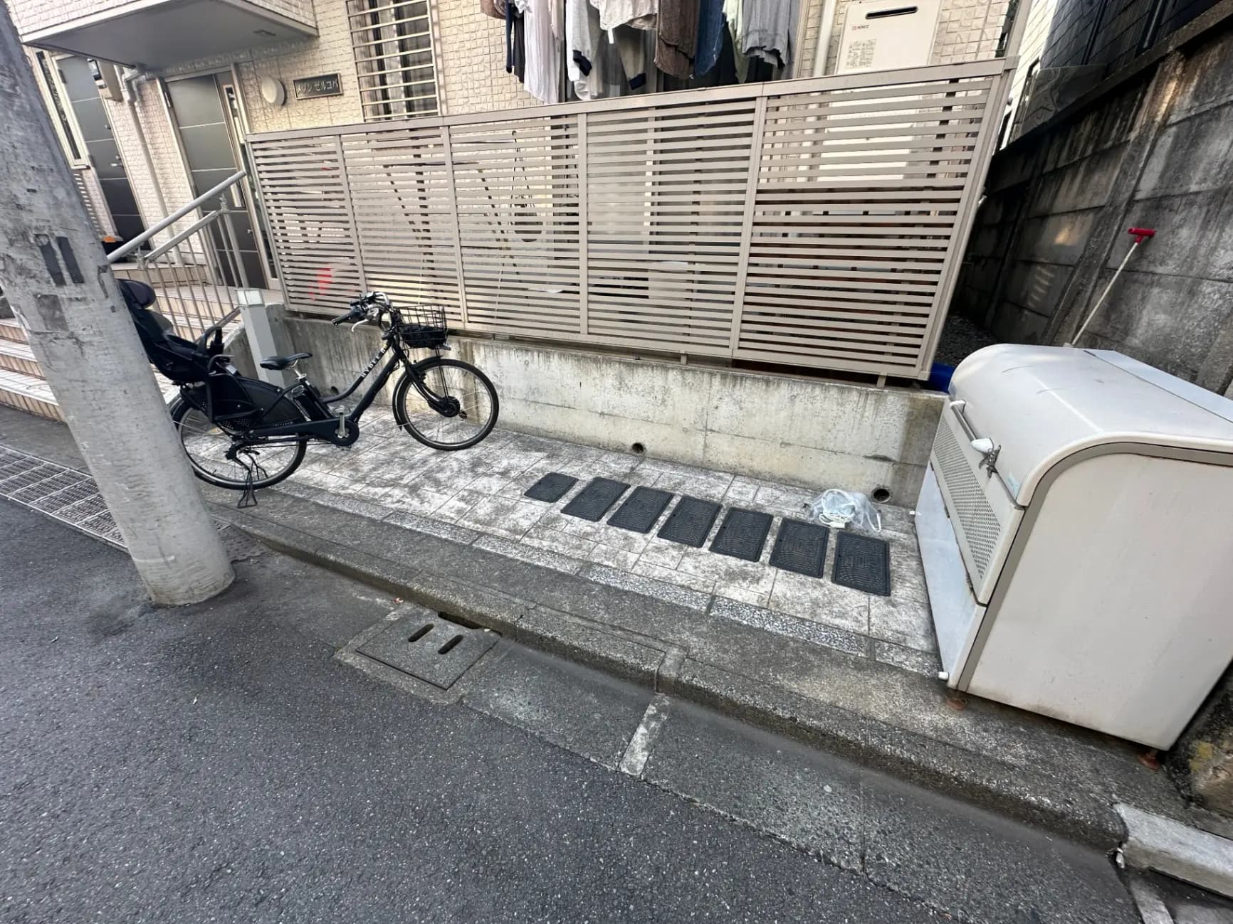 都市部のアパートの外観、駐輪スペースに黒い自転車、エアコン室外機、木製の目隠しフェンス