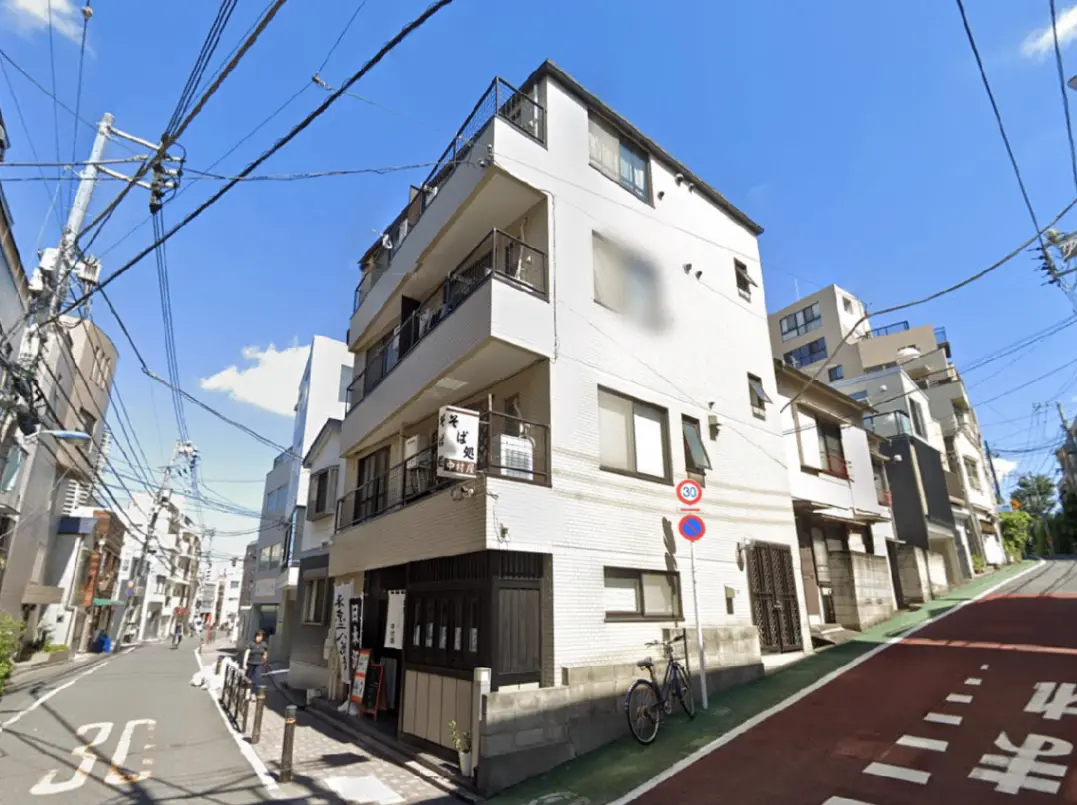 - 白い外壁の四階建てマンション、近くに自転車が置かれた静かな通り、青い空と電柱が映える風景
