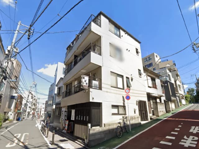 - 白い外壁の四階建てマンション、近くに自転車が置かれた静かな通り、青い空と電柱が映える風景
