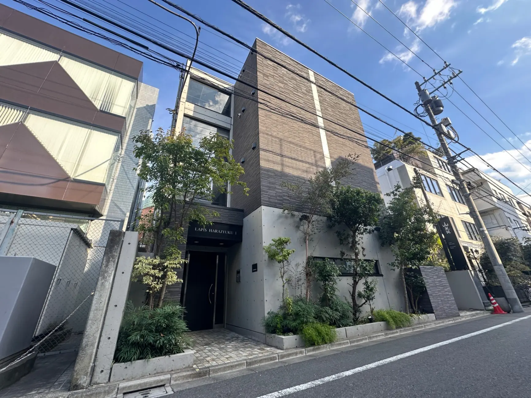 モダンな外観の都市型アパートメント、緑豊かなエントランス、明るい日差しと空が広がる街並み