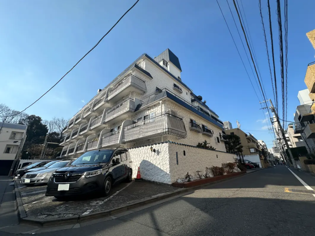 日差しの中にあるホワイトとブルーの外装のマンション、バルコニー付きの多階建て住宅、駐車場に車が並んでいる都市風景