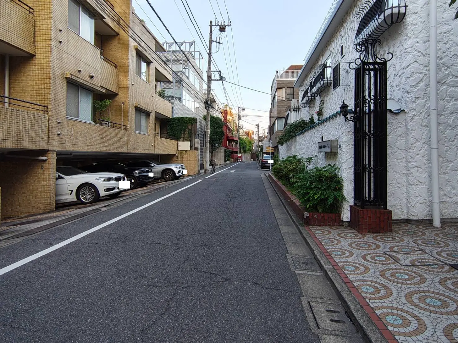 - 都会の住宅街の風景、アパートメントビルの隙間に駐車された車両、石畳の歩道と草花が並ぶエントランスの景観