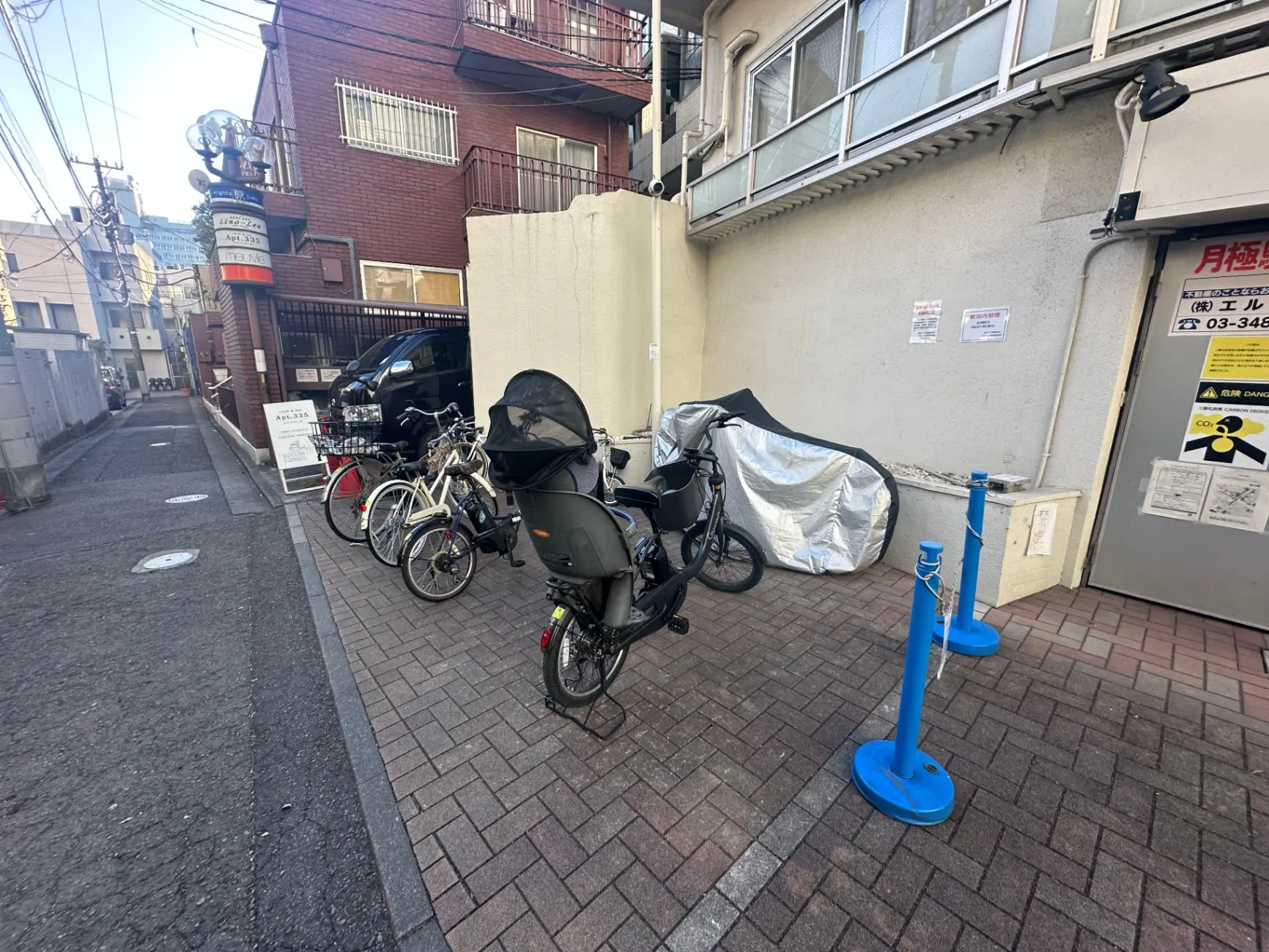 都市の路地に並ぶ自転車とバイク、街角の駐輪場、背景にはレンガの建物と看板