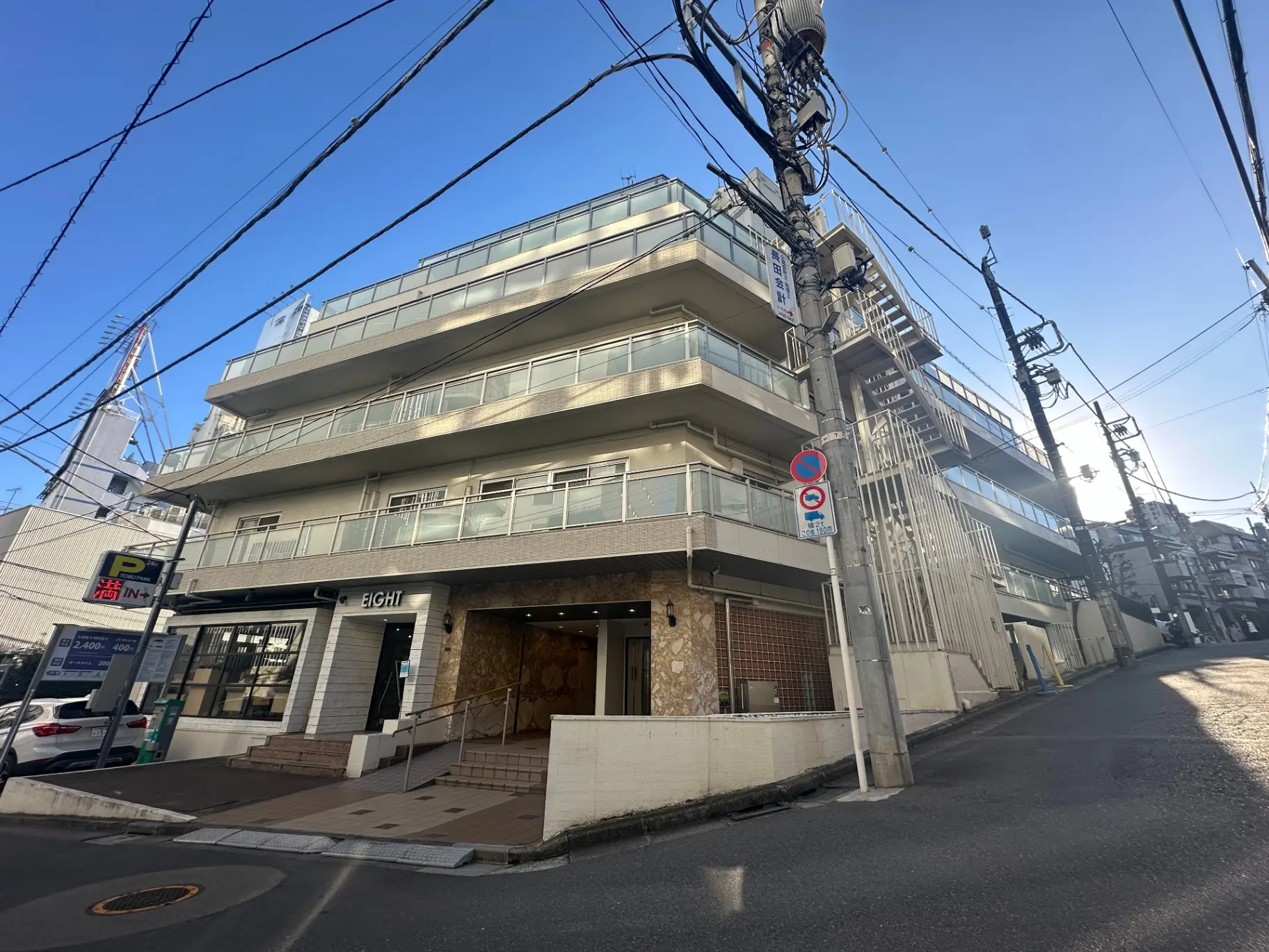 都市の通り沿いに建つモダンな外観の建物、明るい日差しの中、駐車場や看板が見える街角の風景
