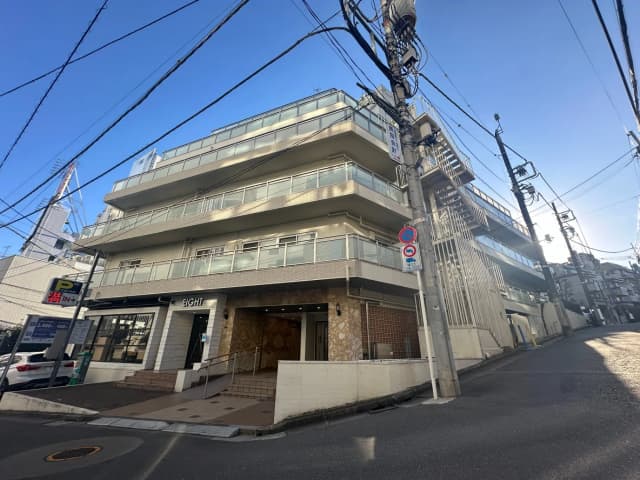 都市の通り沿いに建つモダンな外観の建物、明るい日差しの中、駐車場や看板が見える街角の風景
