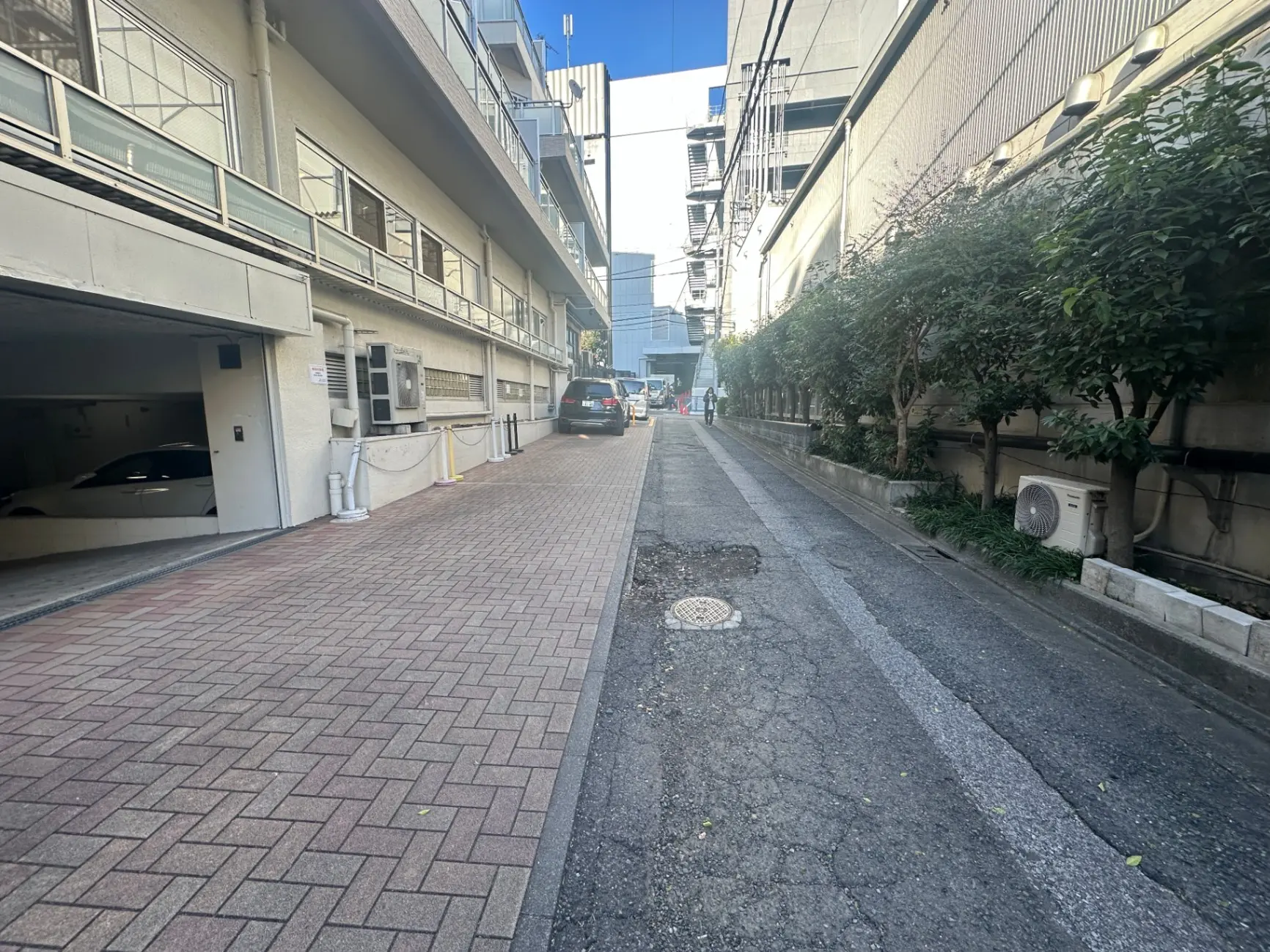 都市のアパートメント街の狭い路地、駐車場入り口と緑の植栽が並ぶ風景