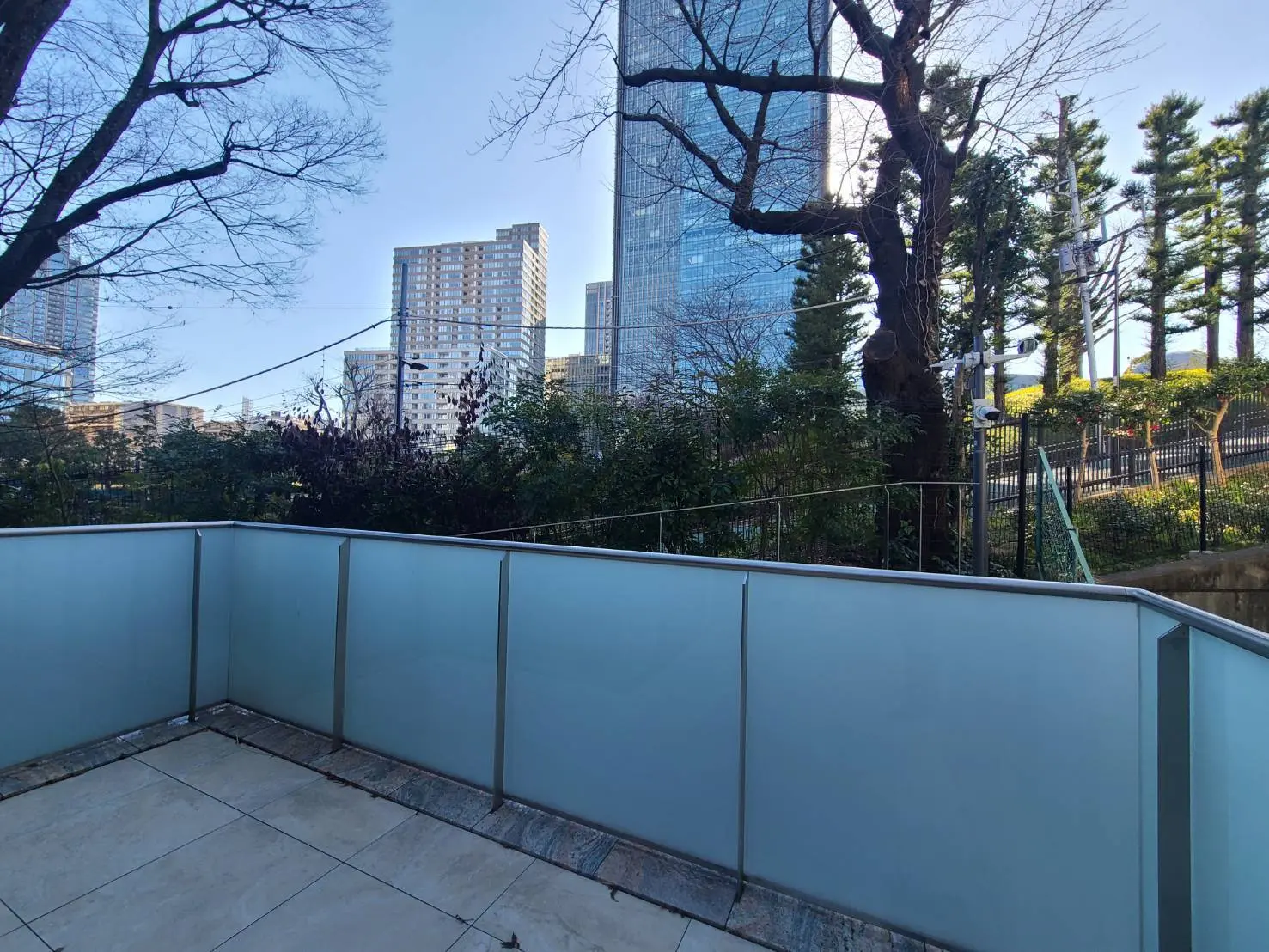 都会の高層ビルと公園の木々が見えるベランダ、青空と都市風景