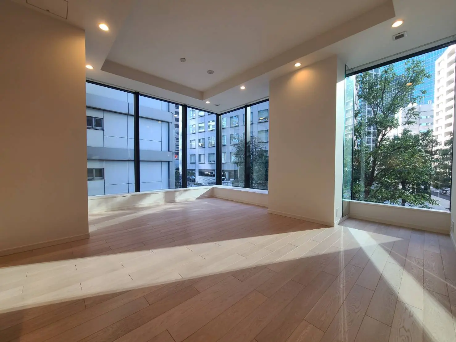 都心のマンションの広々としたリビングルーム、天井からの明るい照明と床から天井までの大きな窓から見える緑の景色