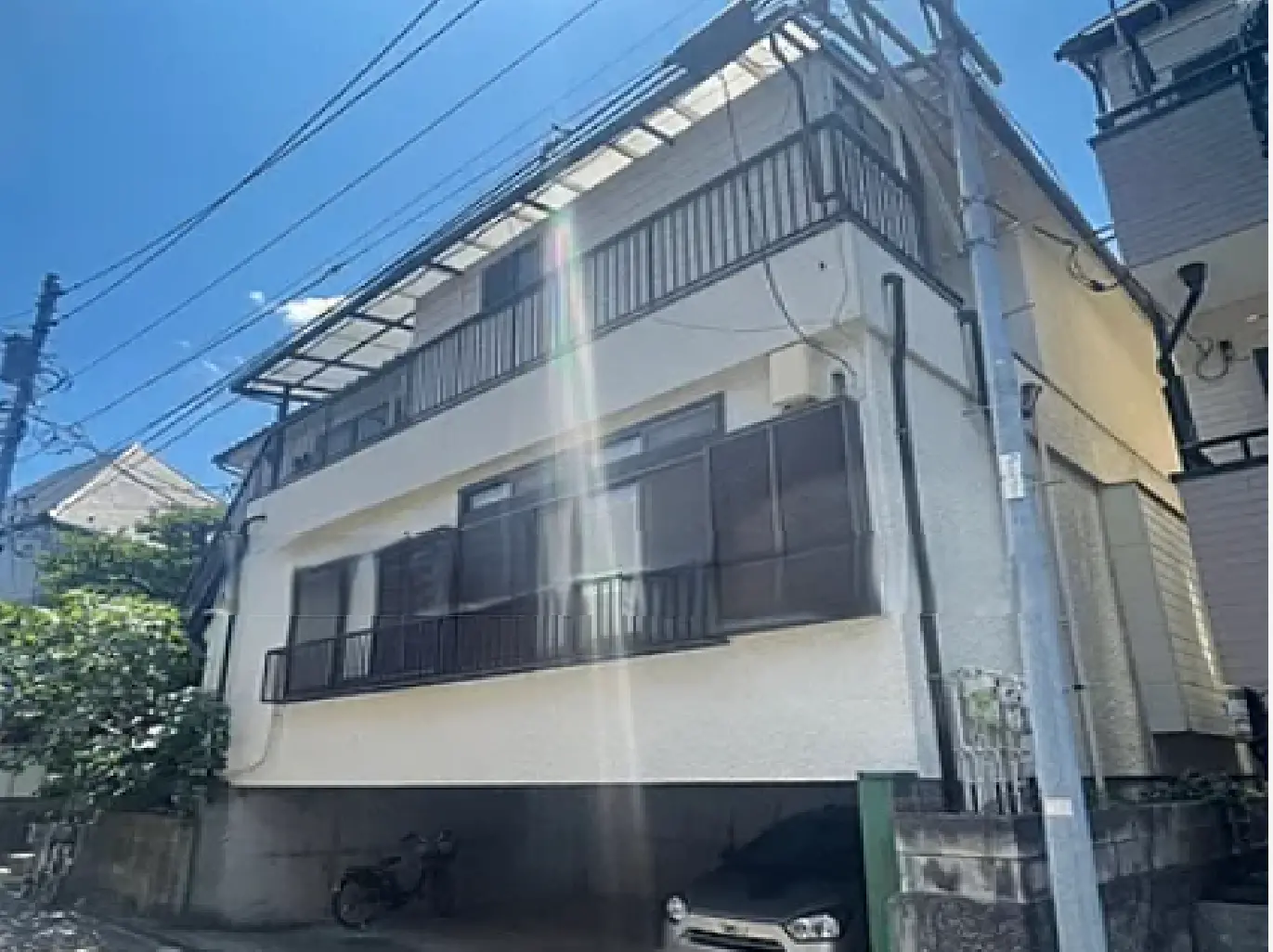 - 白い外壁の三階建て住宅、バルコニーと大きな窓があり、周囲に緑が豊かな住宅街の風景