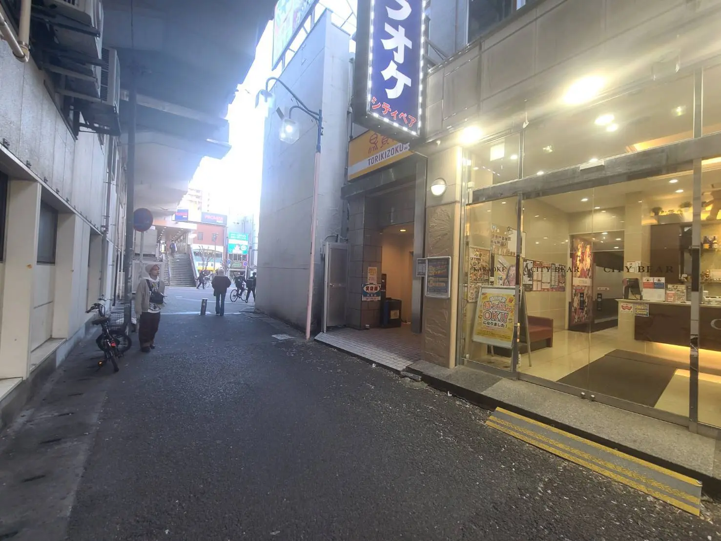 繁華街の裏路地、看板が光る居酒屋と美容院の入口、通りを行く人と自転車