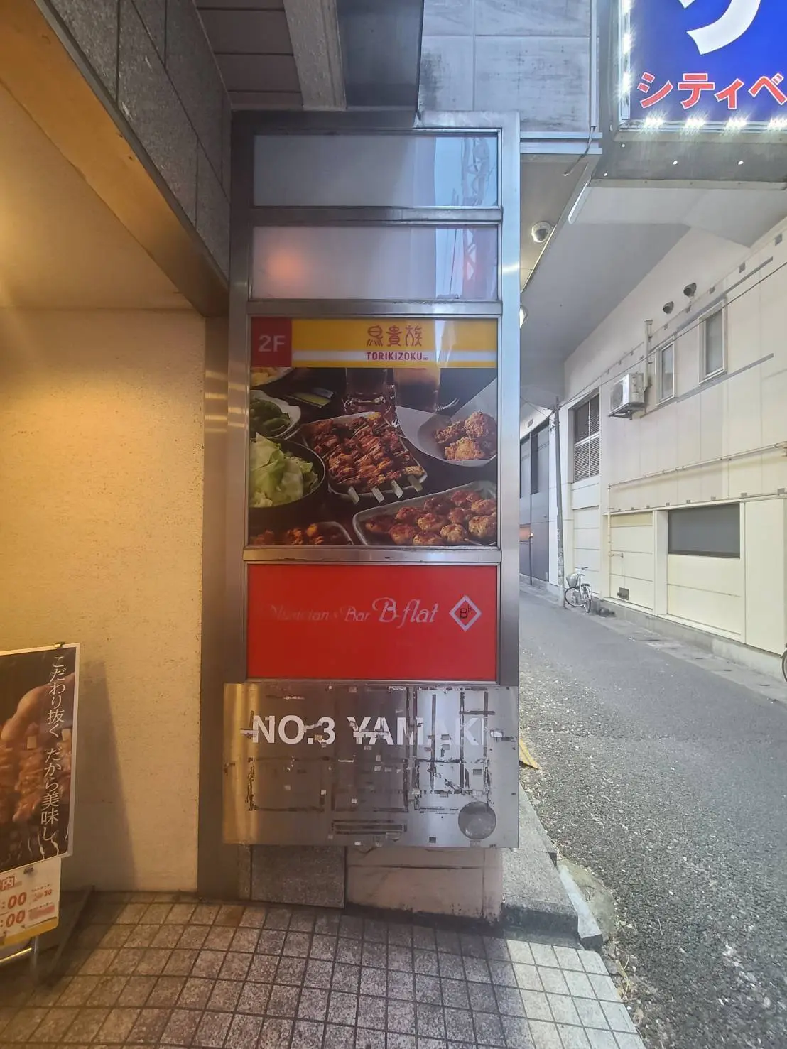 焼き鳥店の看板、居酒屋バーフラットのロゴ、ヤキニクのメニュー写真が掲載された通り沿いのサインボード