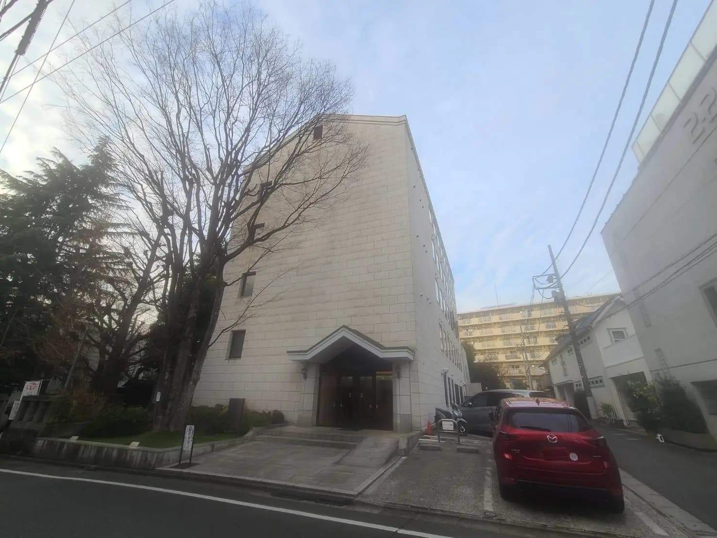 都会の住宅街にあるモダンな外観のマンション、エントランス前に赤い車が駐車されている景観