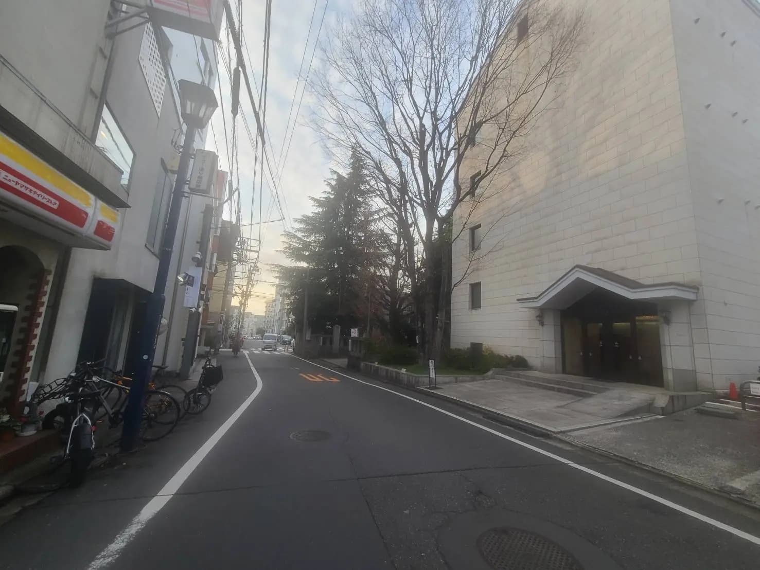 都会の住宅街の道路、左側にコンビニと自転車、右側に建物と木、後方に夕暮れの光が見える風景