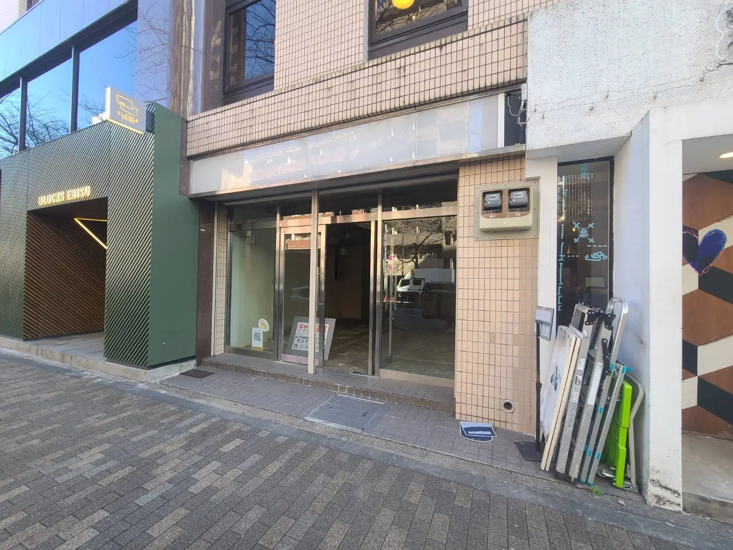 都会のオフィス街にあるモダンなビルの入り口、緑色の建物サインとスキー板が立てかけられている外観