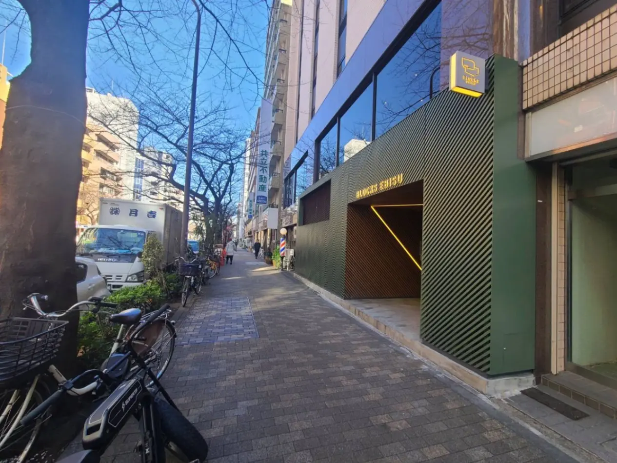 都会の通り沿いにあるモダンなデザインのビル、駐輪スペースと歩道、青空と木々が見える外観