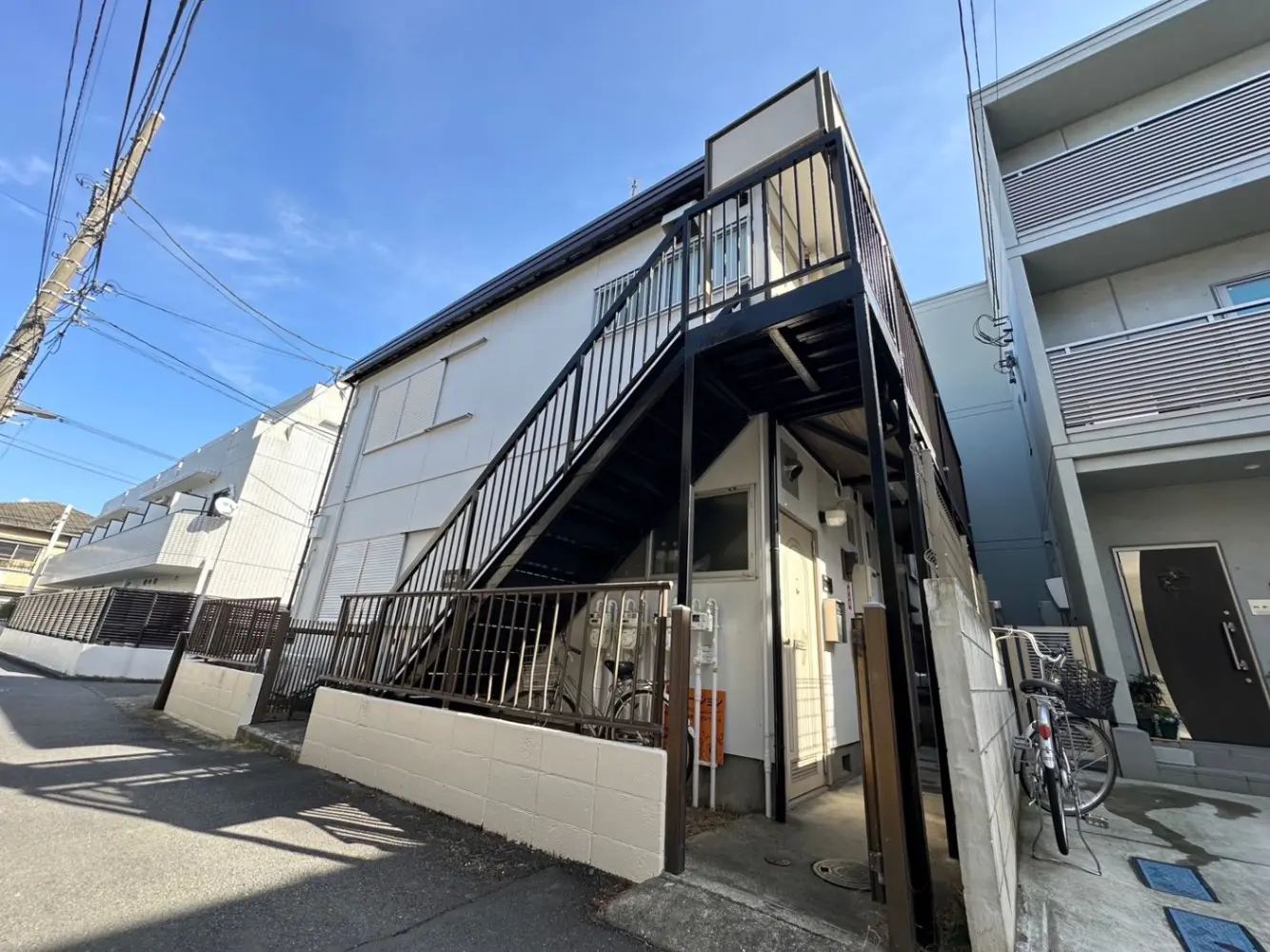 日本の郊外の住宅地にある2階建ての白い外観の家、外階段と駐輪スペース、青空と電線が見える外観