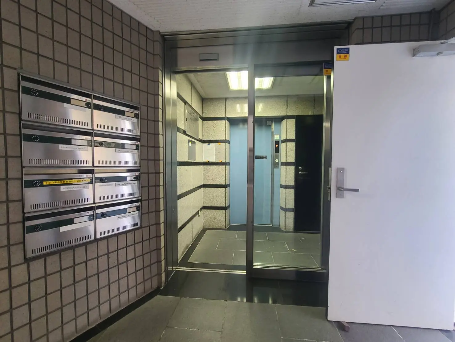 マンションのエントランスホール、インターホン付きの郵便受け、エレベーターと防火扉が見える写真