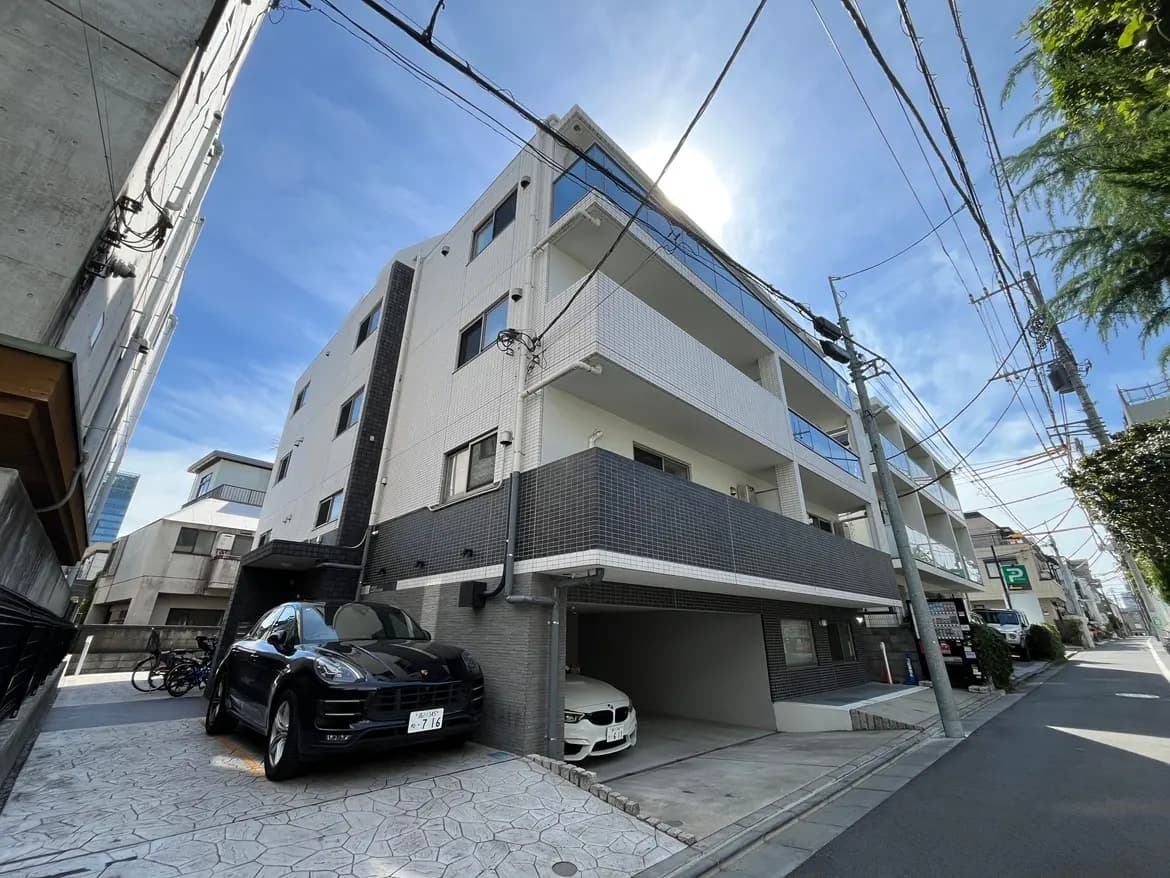 - 現代的な外観のマンション、青空を背景にした白と黒のタイル張りのファサード、駐車場に停まる高級車と自転車が見える景観