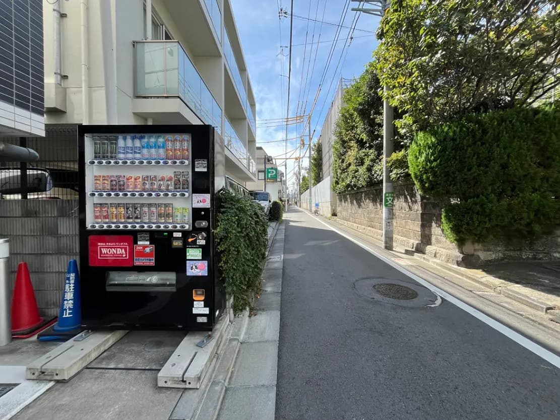 - 道沿いに設置された自動販売機と近隣のマンション、青空と電線が見える日常風景