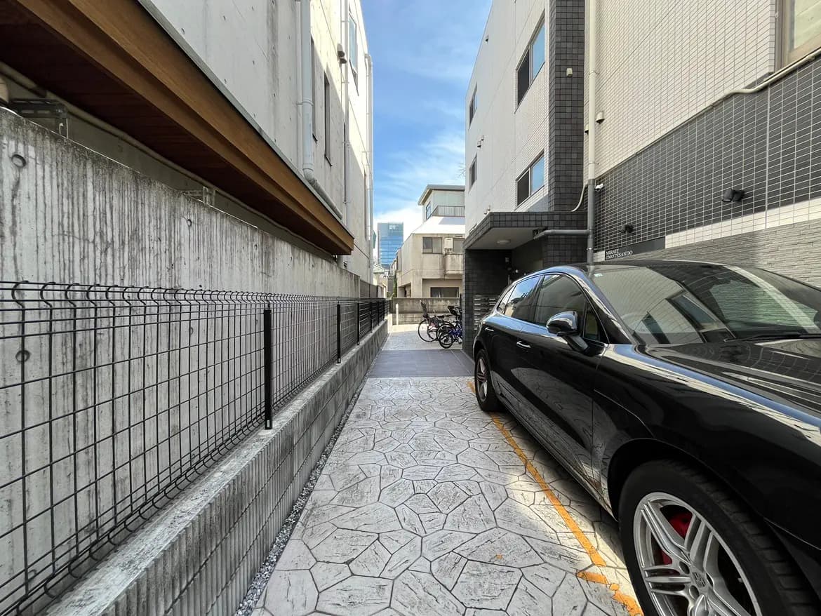 - 近隣のビルに囲まれた狭い通路に駐車された黒い車、コンクリートの壁と石畳の道路が特徴的な風景
