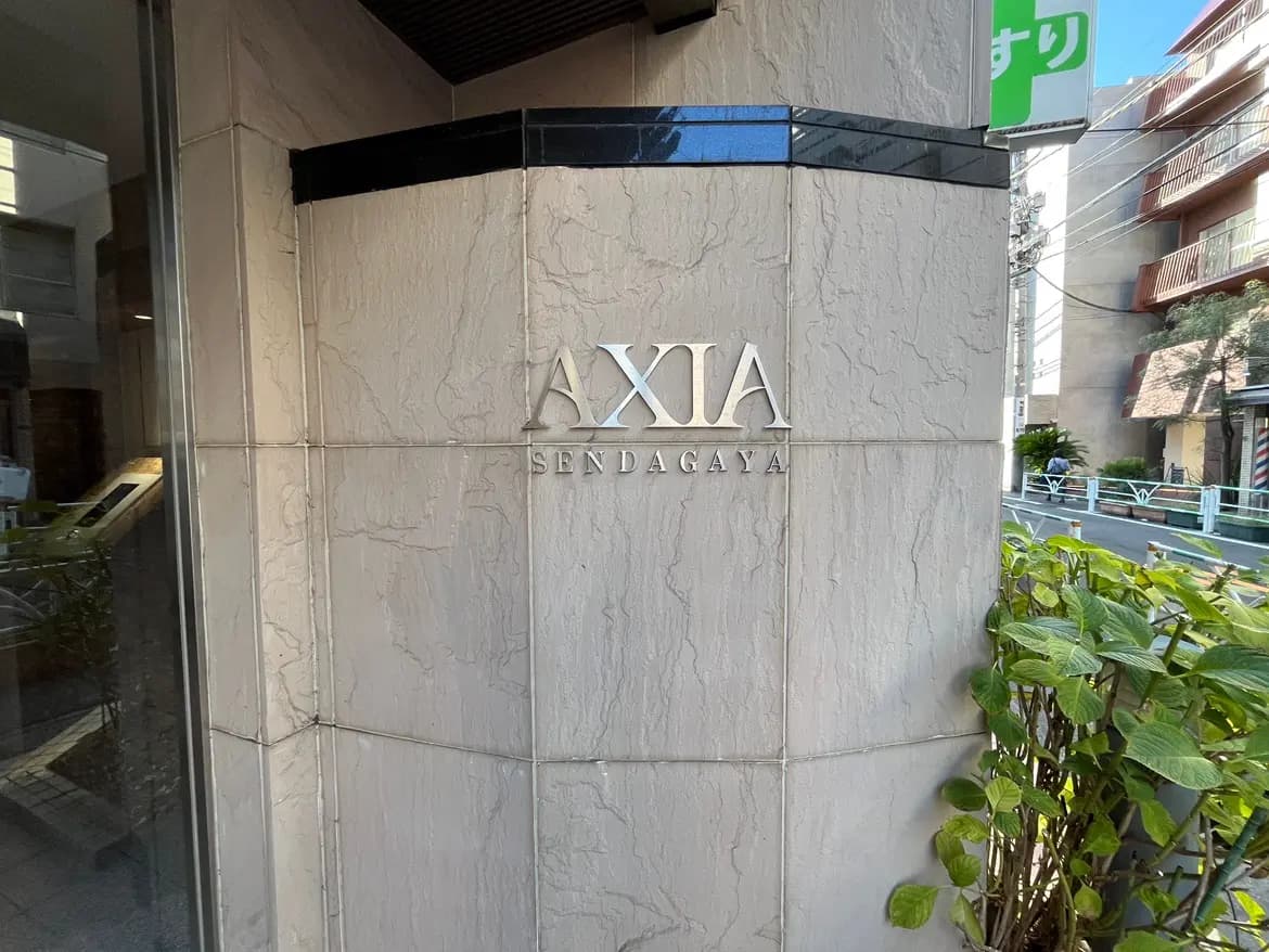 - 「AXIA SENDAGAYA」の看板が設置されたモダンな建物の外観、周囲の緑と街並みが印象的な風景